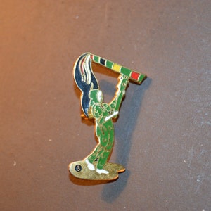 Op de afbeelding: Een gouden speld met een groen figuur dat een vlag vasthoudt met rode, gele en zwarte strepen. Het figuur staat op een gouden voet.