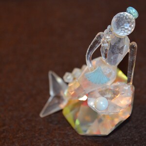 以下が含まれることがあります： クリスタル製の台座に座った透明なガラス製のマーメイドの置物。マーメイドは青い王冠と青い尾びれをしています。台座は虹色で、真珠のようなビーズが付いています。