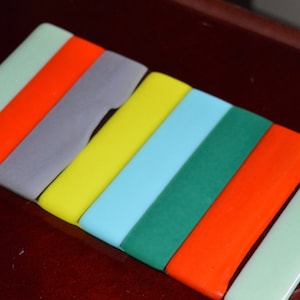 以下が含まれることがあります： 薄い緑、オレンジ、グレー、イエロー、ライトブルー、グリーン、オレンジなど、さまざまな色の長方形のガラスタイル7枚セット。タイルは一列に並べられており、薄い緑のタイルが上、オレンジのタイルが下にあります。