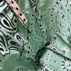 Puede incluir: Un interior de tela verde menta con un patr&oacute;n de bandana de cachemira en blanco y negro. Una correa de tela beige con el mismo estampado de cachemira est&aacute; unida al lateral, terminando en un peque&ntilde;o mosquet&oacute;n de metal color lat&oacute;n antiguo.