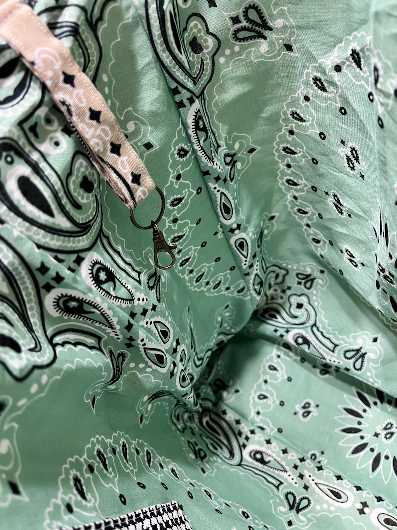 Puede incluir: Un interior de tela verde menta con un patr&oacute;n de bandana de cachemira en blanco y negro. Una correa de tela beige con el mismo estampado de cachemira est&aacute; unida al lateral, terminando en un peque&ntilde;o mosquet&oacute;n de metal color lat&oacute;n antiguo.