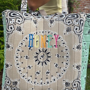 Puede incluir: Un bolso tote acolchado con estampado de bandana color canela, patrones de cachemira negros y costuras verticales. La palabra HONEY est&aacute; bordada en el centro con hilo de colores del arco&iacute;ris. Los paneles laterales son de color verde claro.