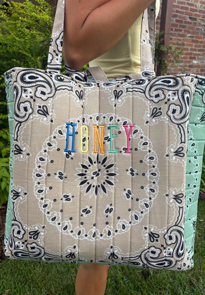 Puede incluir: Un bolso tote acolchado con estampado de bandana color canela, patrones de cachemira negros y costuras verticales. La palabra HONEY est&aacute; bordada en el centro con hilo de colores del arco&iacute;ris. Los paneles laterales son de color verde claro.
