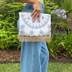 May include: A rectangular white clutch bag featuring a black paisley bandana print pattern. The bag is constructed with vertical quilted stitching and is finished with a row of long, metallic gold fringe along the bottom edge and a gold zipper.