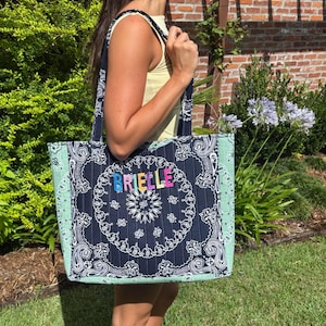 Puede incluir: Un bolso tote acolchado con un panel central de estampado bandana azul marino oscuro con el nombre BRIELLE en letras bordadas de colores. El bolso tiene paneles laterales de color verde menta claro con un patr&oacute;n bandana a juego y dos asas.