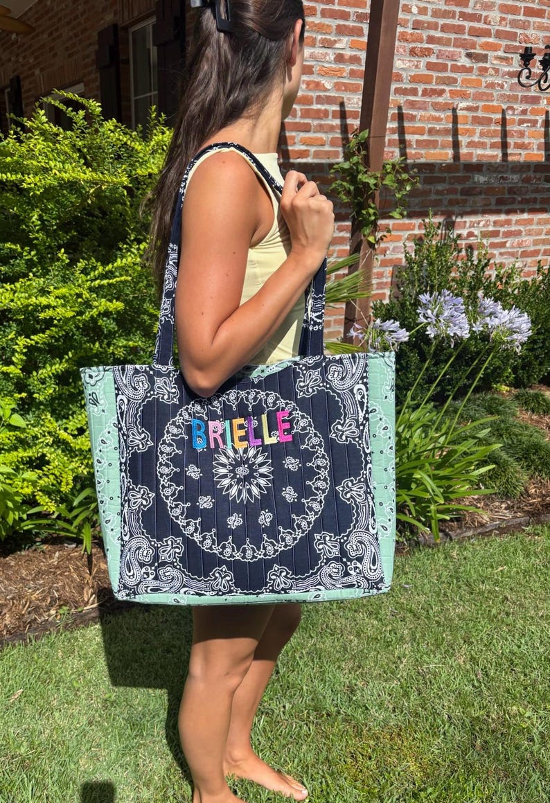 Puede incluir: Un bolso tote acolchado con un panel central de estampado bandana azul marino oscuro con el nombre BRIELLE en letras bordadas de colores. El bolso tiene paneles laterales de color verde menta claro con un patr&oacute;n bandana a juego y dos asas.
