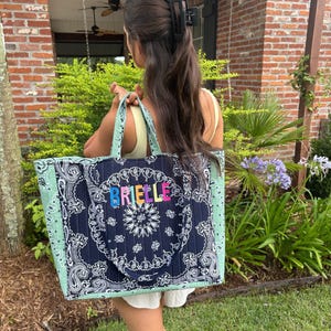 Puede incluir: Un bolso tote acolchado con un patr&oacute;n de bandana de cachemira en azul marino oscuro y blanco en el panel central y cachemira verde salvia claro en los laterales. El nombre BRIELLE est&aacute; bordado en letras coloridas en la parte frontal.