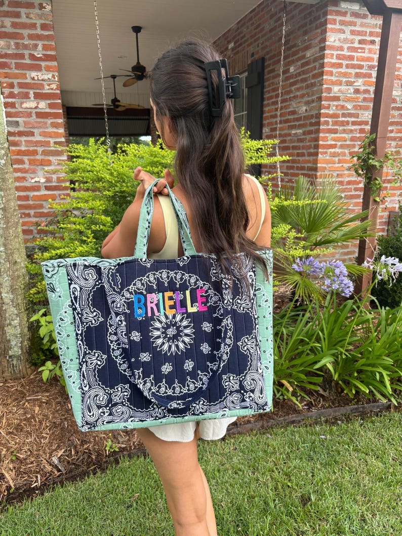 Puede incluir: Un bolso tote acolchado con un patr&oacute;n de bandana de cachemira en azul marino oscuro y blanco en el panel central y cachemira verde salvia claro en los laterales. El nombre BRIELLE est&aacute; bordado en letras coloridas en la parte frontal.