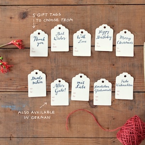 May include: Ten gift tags with handwritten calligraphy in English and German. The tags are white with a scalloped edge and are arranged in two rows on a wooden surface. The tags feature phrases like "Thank you", "Best Wishes", "Happy Birthday", and "Merry Christmas".