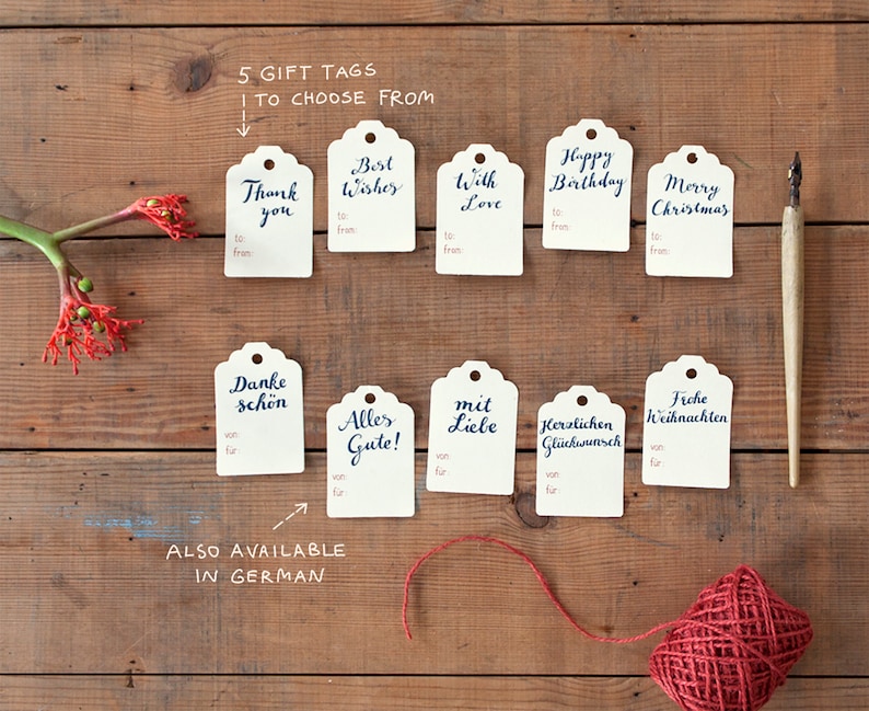 May include: Ten gift tags with handwritten calligraphy in English and German. The tags are white with a scalloped edge and are arranged in two rows on a wooden surface. The tags feature phrases like "Thank you", "Best Wishes", "Happy Birthday", and "Merry Christmas".