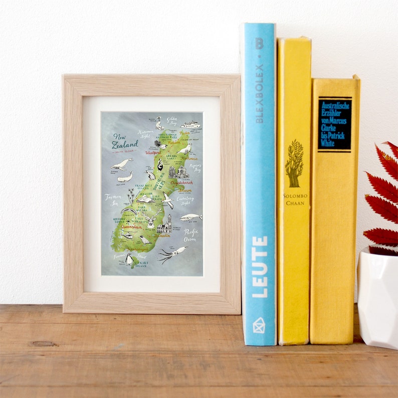 May include: A framed illustration of a map of New Zealand with hand-drawn details of the country's geography, wildlife, and landmarks. The map is in a light wood frame and is sitting on a wooden surface with two books.