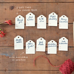 May include: Ten gift tags with handwritten calligraphy in English and German. The tags are white with a scalloped edge and are arranged in two rows on a wooden surface. The tags feature messages such as "Thank you", "Best Wishes", "With Love", "Happy Birthday", "Merry Christmas", "Danke sch&ouml;n", "Alles Gute", "Mit Liebe", "Herzlichen Gl&uuml;ckwunsch", and "Freche Weihnachten".