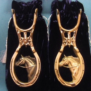 May include: Two gold-colored stirrups with a horse head design inside a horseshoe shape. The stirrups are displayed against a dark blue velvet bag with a silver tassel. Equestrian themed.