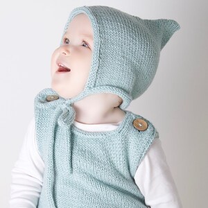 Peut inclure: Un bébé portant un bonnet en tricot bleu clair avec une pointe pointue et un barboteuse sans manches en tricot bleu clair assorti avec des boutons en bois.