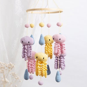 Könnte beinhalten: Ein hängendes Mobile mit gehäkelten Quallen in Pastellgelb, Rosa und Lavendel, durchsetzt mit hellblauen gehäkelten Regentropfen und kleinen Kugeln. Die Figuren sind an einem runden Holzrahmen mit dünnen gelben Schnüren befestigt.