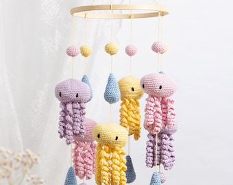 May include: A hanging mobile featuring crocheted jellyfish in pastel yellow, pink, and lavender, interspersed with light blue crocheted raindrops and small spheres. The figures are suspended from a circular wooden frame by thin yellow strings.