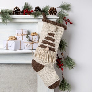 Peut inclure: Une chaussette de Noël en crochet avec un motif d'arbre rayé brun et blanc et une frange blanche. La chaussette est accrochée à une cheminée blanche avec quelques petits cadeaux emballés.
