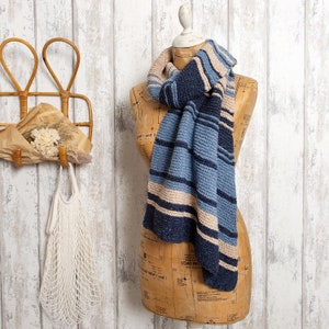 Può includere: Una sciarpa a righe blu e beige con un motivo a maglia testurizzato. La sciarpa è drappeggiata su un manichino con uno sfondo bianco.