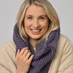 Könnte beinhalten: Eine Frau trägt einen lilafarbenen Strick-Infinity-Schal. Der Schal ist dick und grob gestrickt, und die Frau lächelt.