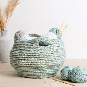 May include: A round, pale blue and cream-coloured woven basket filled with balls of yarn and wooden crochet hooks. The basket has two handles and is on a neutral surface. More yarn and a hook are beside the basket.