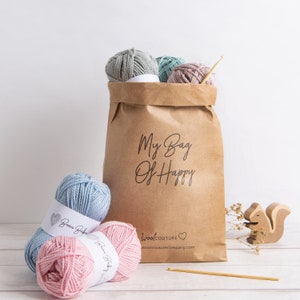 May include: A brown paper bag with the text "My Bag Of Happy" printed on it. The bag is filled with balls of yarn in shades of blue, green, and pink. The yarn is labeled "Beau Baby" and "Wool Couture". A wooden squirrel figurine and a crochet hook are also in the image.