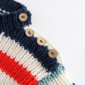May include: Close-up of a striped knitted jumper with a white, red, blue and green colour scheme. The jumper has wooden buttons.