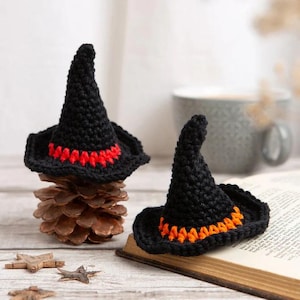 Può includere: Due cappelli da strega in crochet nero con bordi arancioni e rossi. I cappelli sono appoggiati su una pigna e un libro.