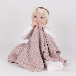 May include: A baby wearing a white knitted headband with a button detail is sitting on a white background and holding a pink knitted blanket.