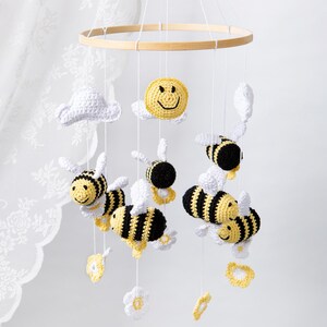 May include: A handmade baby mobile featuring crocheted bees with yellow and black stripes, white wings, and smiling faces. The mobile also includes white clouds, yellow flowers, and a smiling sun. The elements are suspended from a wooden hoop.
