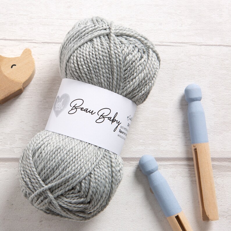 May include: A skein of light gray yarn labeled "Beau Baby" with a white label and a heart logo. The yarn is on a white wooden surface with two blue painted wooden clothespins.