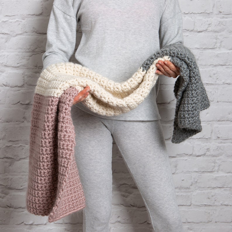 May include: A crocheted blanket with three sections in pink, cream, and gray. The blanket is folded and held by a person.