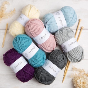 May include: A selection of colourful yarn skeins in shades of cream, pink, light blue, teal, grey, purple, and charcoal. Each skein has a white label with the text "Beautifully Basic". Wooden knitting needles are also visible.