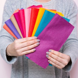 May include: A collection of felt fabric swatches in various colours, including purple, teal, yellow, orange, red, pink, and brown. The swatches are fanned out and held in a person's hands.