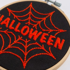 May include: Embroidered Halloween decoration with a black fabric background and orange thread. The word "Halloween" is stitched in a bold font with a spiderweb design behind it.