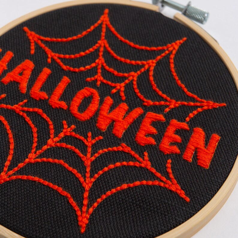 May include: Embroidered Halloween decoration with a black fabric background and orange thread. The word "Halloween" is stitched in a bold font with a spiderweb design behind it.