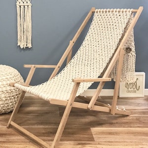 May include: A white macrame chair with a wooden frame. The chair is folded and has a white macrame pillow on the floor next to it. The chair is in front of a blue wall.