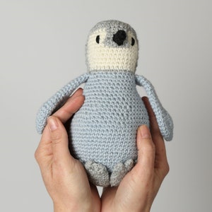 May include: A crocheted penguin toy with a light blue body, white belly, and black eyes. The penguin is being held by two hands.