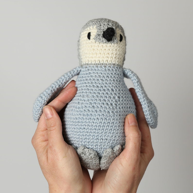 May include: A crocheted penguin toy with a light blue body, white belly, and black eyes. The penguin is being held by two hands.