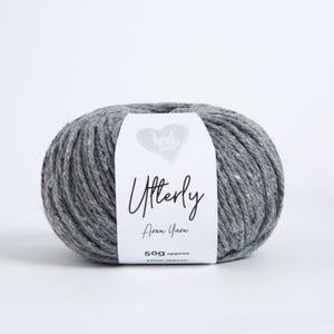 K&ouml;nnte beinhalten: Ein Kn&auml;uel graues, gesprenkeltes Aran-Garn mit einem wei&szlig;en Papieretikett, das ein Herzsymbol, den Text Wool Couture, Utterly, Aran Yarn und das aufgedruckte Gewicht von 50g approx. zeigt. Das Garn hat eine strukturierte Optik mit kleinen wei&szlig;en Flecken.