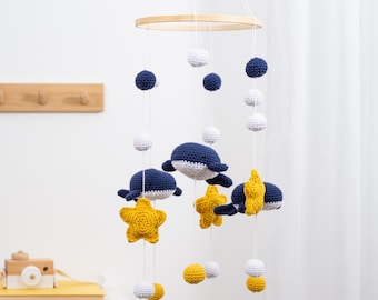 May include: A hanging nursery mobile featuring crocheted navy blue whales and yellow stars suspended from a circular wooden frame. The mobile includes white, navy blue, and yellow crocheted spheres attached to white strings, creating a layered design.