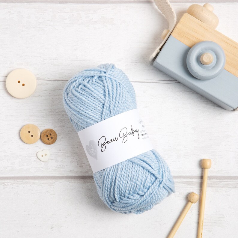 May include: A light blue ball of yarn with a white label that says "Beau Baby" on it. The yarn is on a white wooden surface with two wooden knitting needles and some buttons.