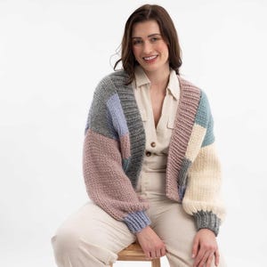 May include: A chunky knit cardigan in a patchwork design. The sweater features blocks of gray, lavender, pink, blue, and cream colors. The cardigan has long sleeves and a relaxed fit, perfect for layering. The model is wearing a beige button-down shirt and beige pants.