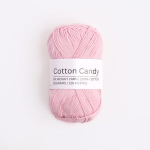 May include: A skein of light pink DK weight yarn made of 100% cotton. A white paper label wraps around the center, printed with the text Cotton Candy, DK WEIGHT YARN | 100% COTTON, and 50GRAMS / 125 METRES in black sans-serif lettering.