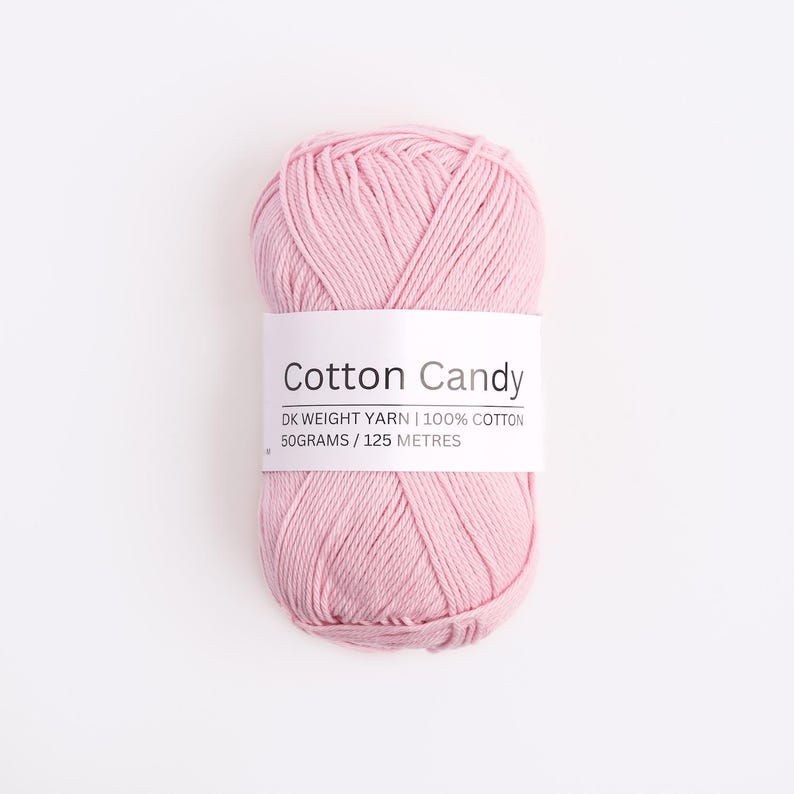 May include: A skein of light pink DK weight yarn made of 100% cotton. A white paper label wraps around the center, printed with the text Cotton Candy, DK WEIGHT YARN | 100% COTTON, and 50GRAMS / 125 METRES in black sans-serif lettering.