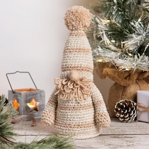 May include: A crocheted gnome with a striped hat and a brown beard. The gnome is standing on a wooden surface with a Christmas tree and other decorations in the background.