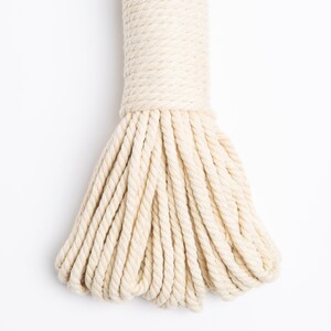 May include: A close-up of a thick, natural cream-colored cotton rope. The rope is tightly wound and has a soft, textured appearance.