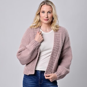 May include: A dusty rose-colored, chunky knit cardigan with a ribbed texture. The open-front sweater has long sleeves and a relaxed fit. The model is wearing a white t-shirt and blue jeans. The cardigan is a cozy and stylish outerwear option.