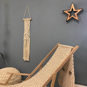 May include: A beige macrame chair with a fringe detail and a wooden frame. The chair is in front of a gray wall with a beige macrame wall hanging and a wooden star with lights.