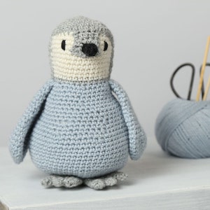May include: A crocheted penguin toy with a gray head, white face, and light blue body. The penguin is standing on a white surface.