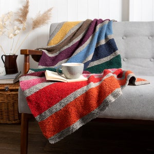 May include: A colorful striped knitted throw blanket draped over a gray armchair with a mug of coffee and a book on top.